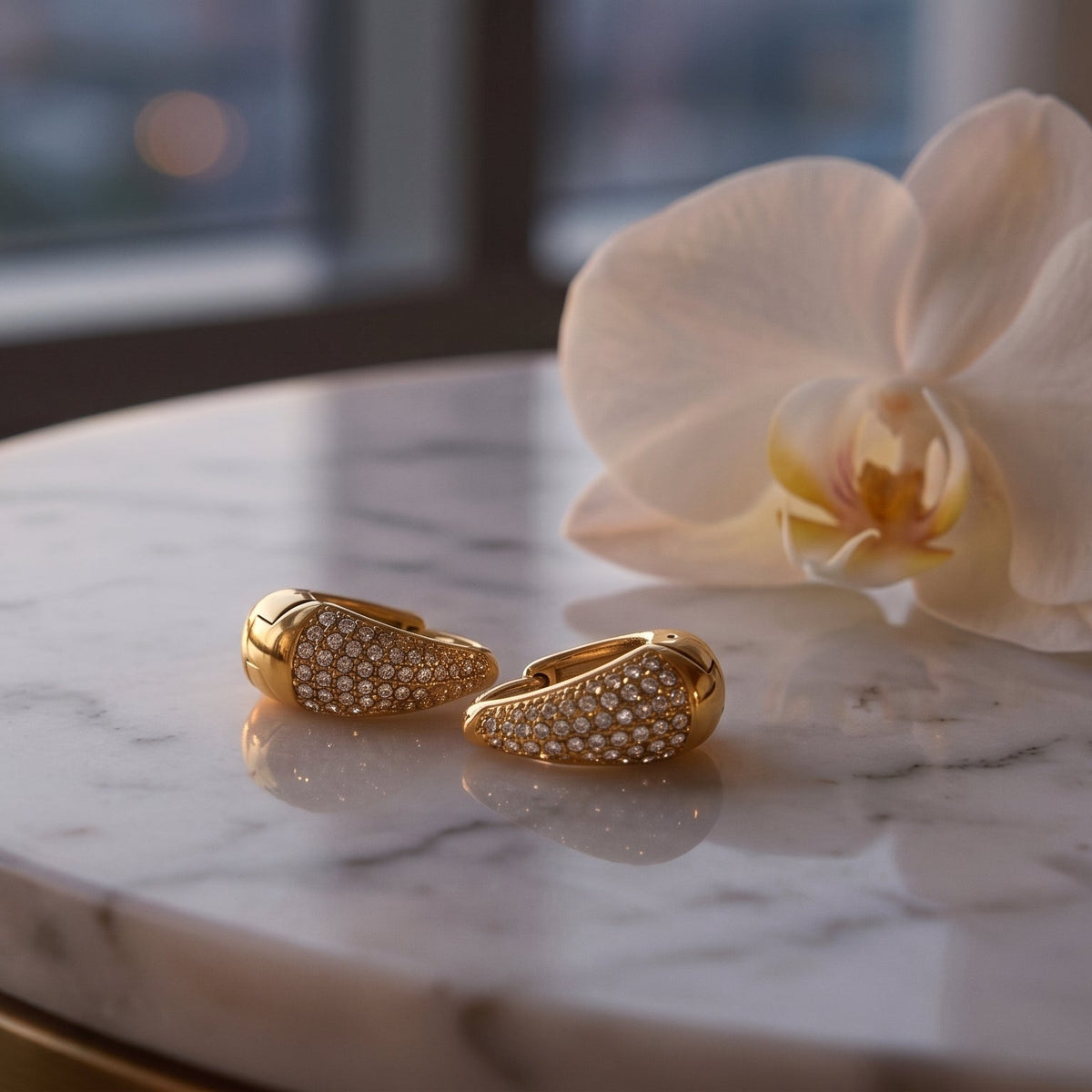 A pair of gold rings with pavé-set clear stones resting on a white marble table next to a white orchid flower, with a softly blurred window background.