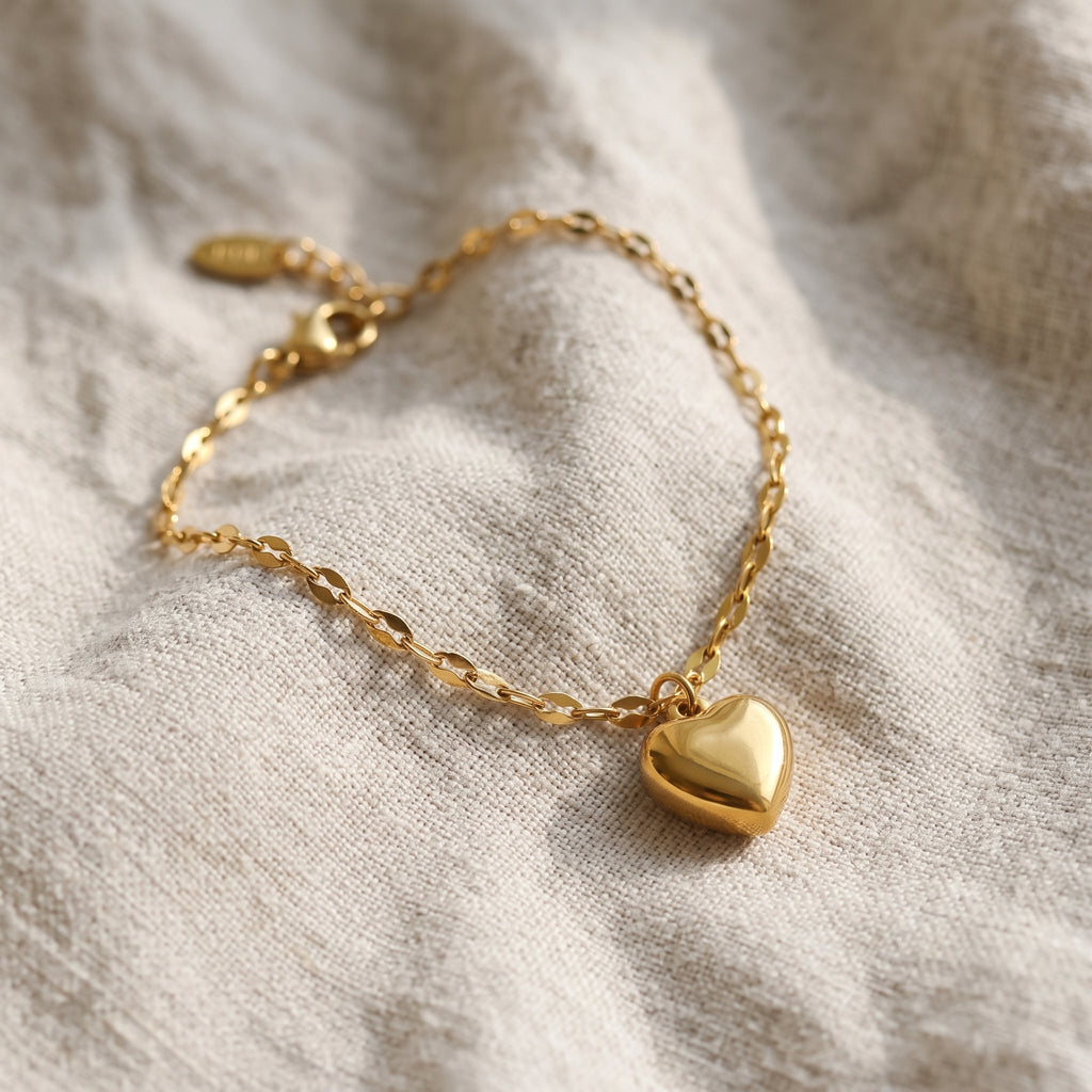 A delicate gold chain bracelet with a polished heart-shaped pendant, resting on a textured beige linen fabric.