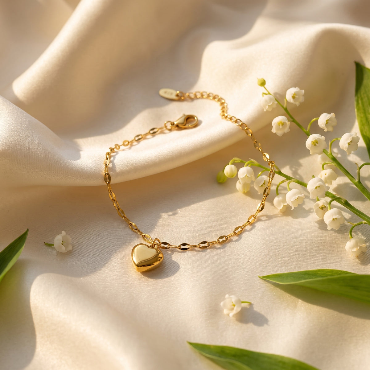 A delicate gold chain bracelet with a polished heart charm, styled on draped cream satin fabric with white lily of the valley flowers and green leaves.