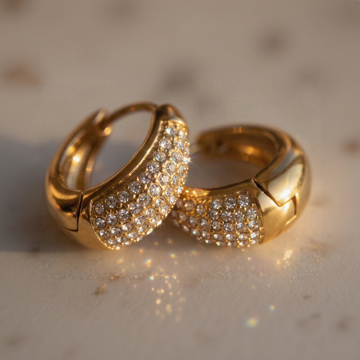 Two gold hoop earrings with rows of clear pavé gemstones, resting on a light marble surface under warm, direct lighting.