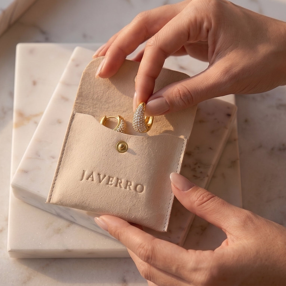 A close-up of hands holding a beige velvet jewelry pouch embossed with the brand name 'JAVERRO', which is open to reveal a gold ring inside, set on a white marble surface.