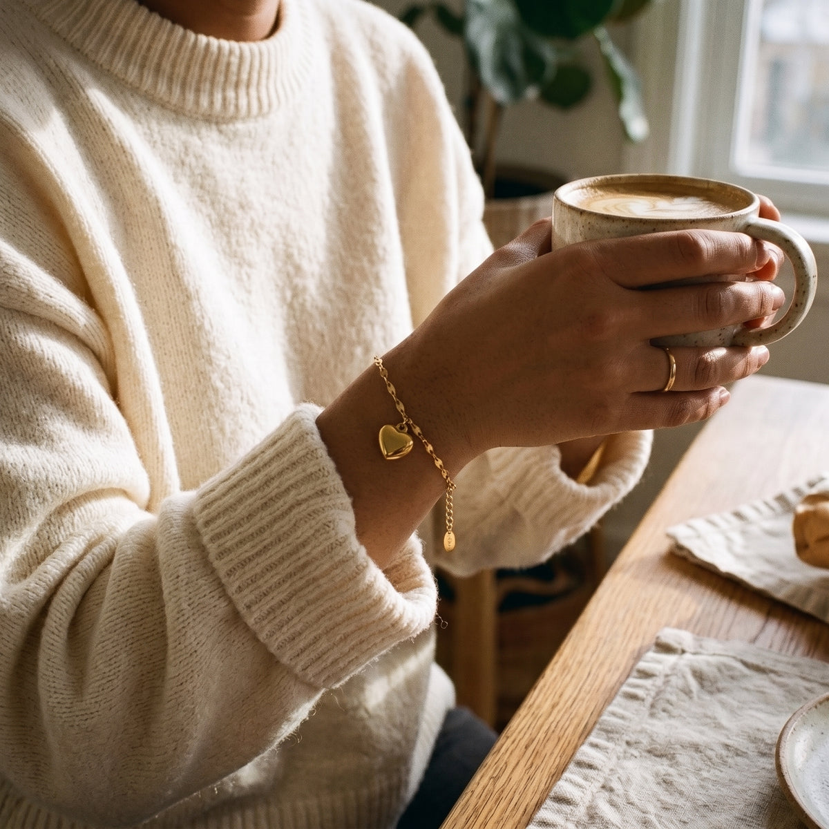 A women in a cream knit sweater holds a ceramic mug at a wooden table, wearing a delicate gold chain bracelet with a small heart charm.